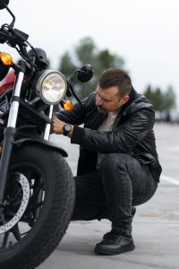 Um homem de jaqueta de couro preta e calça jeans, agachado ao lado de uma moto vermelha, inspeciona ou ajusta a parte frontal da moto. Ele está ao ar livre, em um estacionamento.