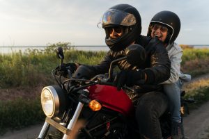 Um homem e uma mulher, ambos usando capacetes, em uma moto vermelha, rodando em uma estrada de terra. A mulher, sentada atrás, está sorrindo.