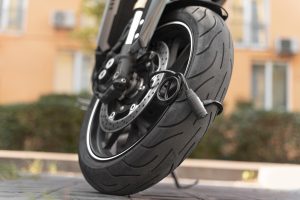 Pneu dianteiro de uma moto, com um cadeado em disco preso ao freio a disco, garantindo segurança. A banda de rodagem do pneu tem um desenho que sugere uso em asfalto, e o contexto da imagem é urbano, com um prédio ao fundo.