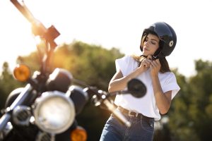 Uma mulher de camiseta branca e calça jeans, em frente a uma moto, está ajustando a fivela de seu capacete preto. O sol brilha atrás dela.