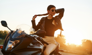 Uma mulher jovem e elegante sentada em uma motocicleta esportiva preta durante o pôr do sol. Ela veste uma jaqueta de couro preta aberta, blusa preta e calças beges, usando óculos de sol redondos enquanto ajusta o cabelo em um rabo de cavalo. A luz quente do sol cria um efeito de brilho intenso ao fundo, destacando a silhueta da motociclista e da moto.